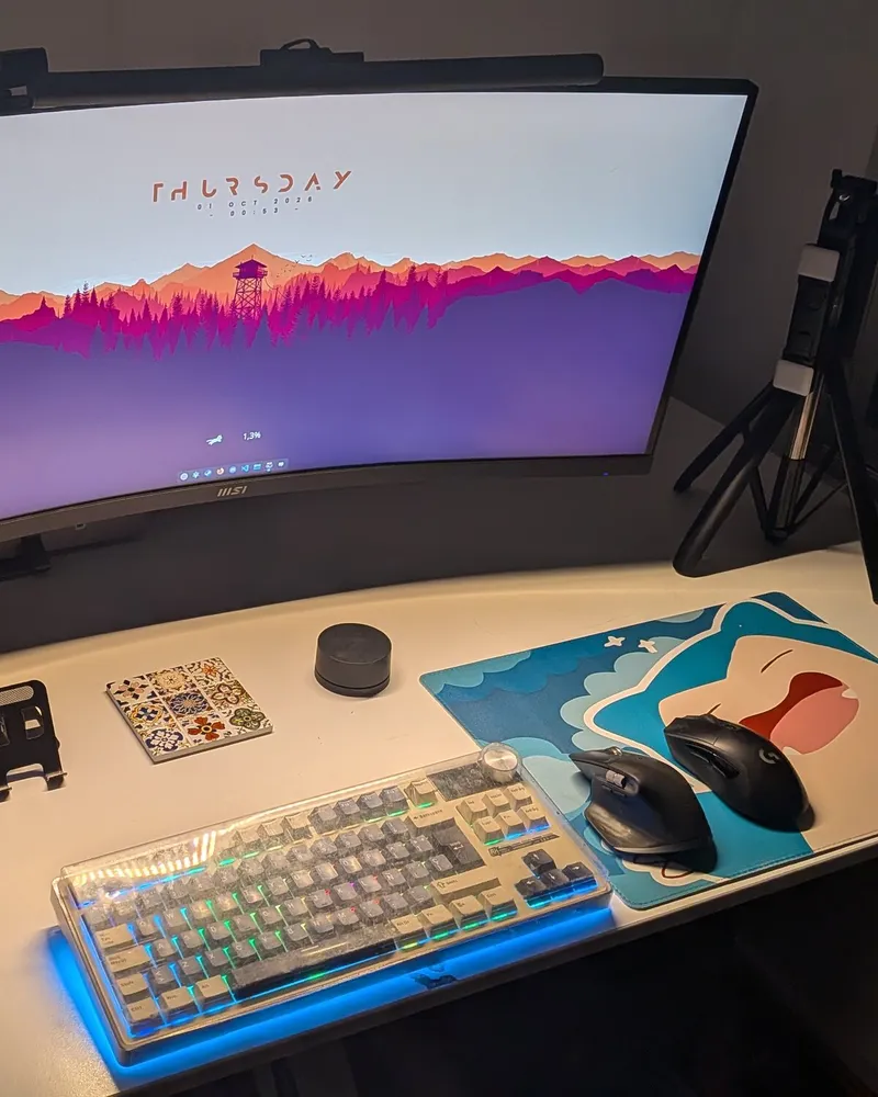 My desk in 2026: a curved ultrawide monitor with the mountain wallpaper, a transparent mechanical keyboard with blue lighting, two mice and a Snorlax desk mat.